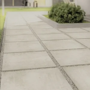 A neatly laid stone pathway featuring the Latte Outdoor Porcelain Paving Tiles – 600mm x 900mm x 20mm, surrounded by small gravel, leads through a garden. In the background, there is green grass and a bushy plant near a building.