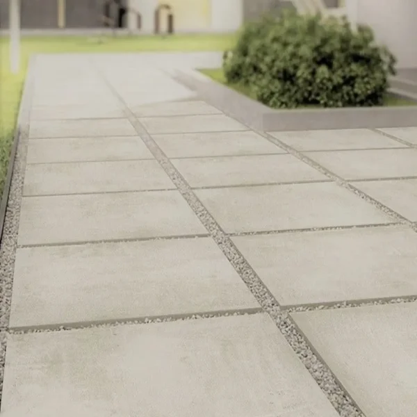 A neatly laid stone pathway featuring the Latte Outdoor Porcelain Paving Tiles – 600mm x 900mm x 20mm, surrounded by small gravel, leads through a garden. In the background, there is green grass and a bushy plant near a building.