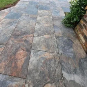 Rustic Slate Outdoor Porcelain Paving Tiles (600mm x 900mm x 20mm) feature a natural stone look in gray, brown, and blue shades, laid in a staggered pattern beside green plants and a brick wall.
