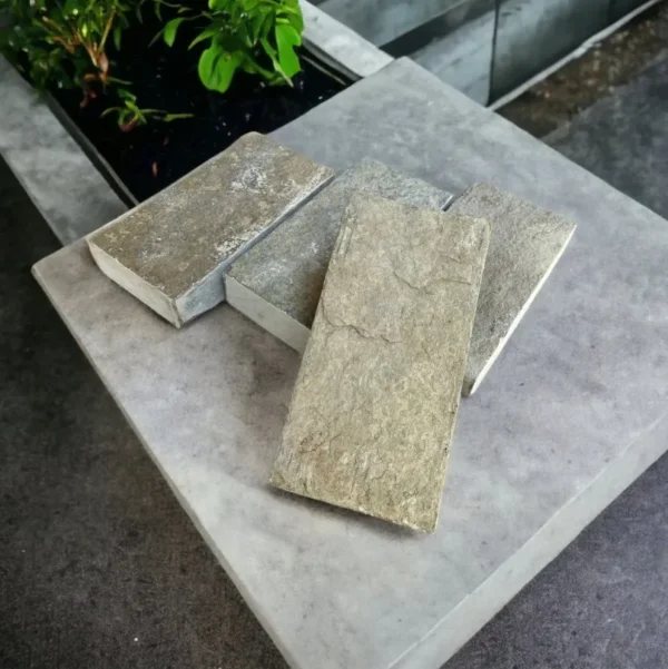 Four rectangular Yellow Limestone Cobble Setts Sawn And Tumbled 100x200x40mm are arranged overlapping on a smooth concrete surface near a planter with green leafy plants.