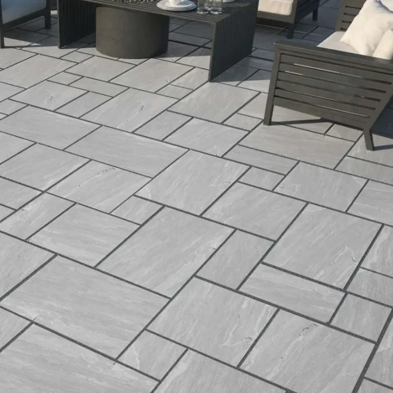A patio features Kandla Grey Outdoor Porcelain Paving Tiles in mixed sizes arranged in a geometric, interlocking pattern, paired with modern outdoor furniture with dark frames and light cushions.