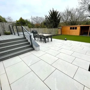 A modern backyard features a raised deck with steps, a dining set on Himalayan White Outdoor Porcelain Paving Tiles (600mm x 900mm x 20mm), artificial grass, a wooden shed, and a tall fence with trees in the background.