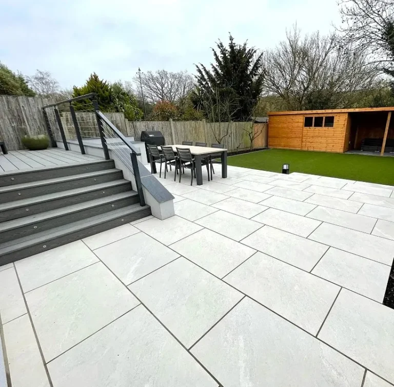 A modern backyard features a raised deck with steps, a dining set on Himalayan White Outdoor Porcelain Paving Tiles (600mm x 900mm x 20mm), artificial grass, a wooden shed, and a tall fence with trees in the background.