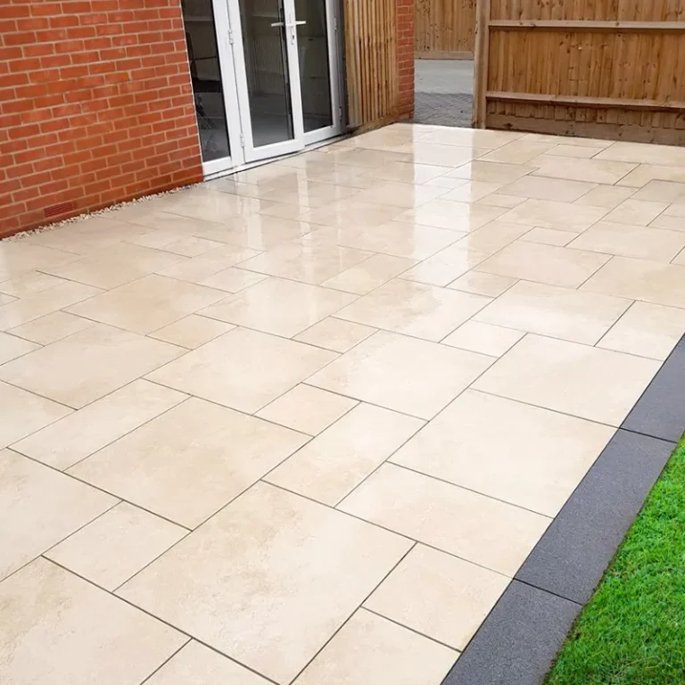 A patio paved with HS Beige Outdoor Porcelain Paving Tiles – Mixed Sizes sits outside a brick building with glass doors, bordered by a wooden fence and green grass.