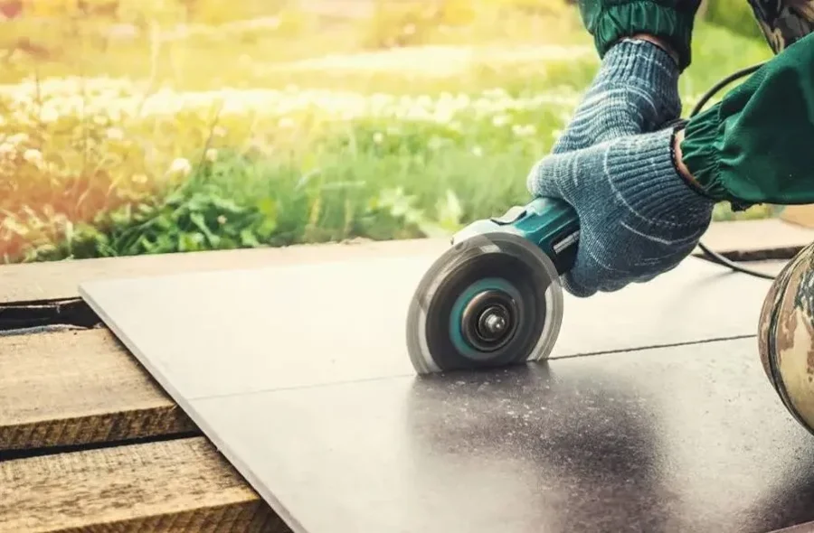 A person wearing gloves and a green jacket uses an electric angle grinder to cut a dark tile placed on a wooden surface outdoors.