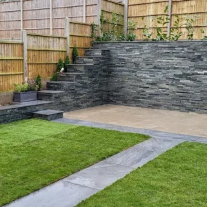 A modern backyard with Silver Grey Stone Cladding on the patio and steps, tiered garden beds, wooden fencing, and neatly trimmed grass sections separated by a slate pathway.