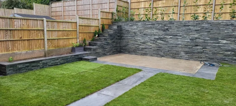 A modern backyard with Silver Grey Stone Cladding on the patio and steps, tiered garden beds, wooden fencing, and neatly trimmed grass sections separated by a slate pathway.