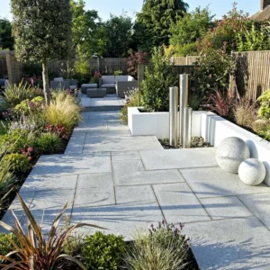 Modern garden with Silver Grey Granite Paving Slabs (mixed sizes, 20mm), ornamental grasses, shrubs, a metal water feature with tall pipes and two large stone spheres in the foreground, and a gray outdoor seating area in the background.