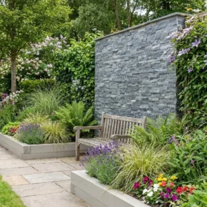 A wooden bench sits on a stone patio beside vibrant flower beds. Behind the bench, a tall feature wall clad in Silver Shine Cladding is surrounded by lush green foliage and climbing plants.