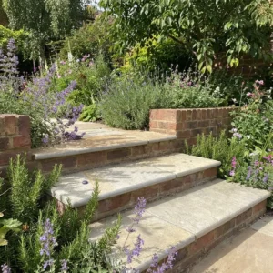 Yellow Limestone Bullnose Edging 900x350x35mm lines the stone steps leading through a lush garden filled with blooming purple, pink, and green plants, surrounded by vibrant foliage and trees on a sunny day.
