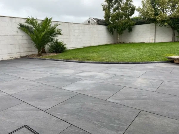 A backyard featuring a spacious patio adorned with County Gris Outdoor Porcelain Paving Tiles, measuring 600mm x 900mm x 20mm, and a small grassy section. Several small trees and plants are neatly positioned along the white brick wall that encircles the garden. The sky is overcast.
