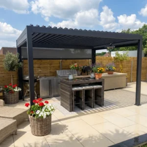 A modern outdoor kitchen under a black pergola with a bar counter, stools, stainless steel appliances, wicker furniture, and potted plants sits on an Aosta Creama porcelain 600 x 900 tiled patio beside a wooden privacy fence under a partly cloudy sky.