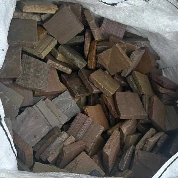 A close-up view of a bag filled with Autumn Brown Sandstone Cobble Setts (100mm x 100mm x 22mm, Handcut Edge) in brown, gray, and beige shades. The textured stones are stacked unevenly.