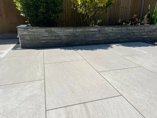 A close-up view of a modern garden patio showcases large rectangular tiles crafted from Everest Pearl Outdoor Porcelain Paving Tiles 600mm x 900mm. In the background, a raised garden bed made with gray brick features green shrubs and a wooden fence. Sunlight casts gentle shadows across the scene.