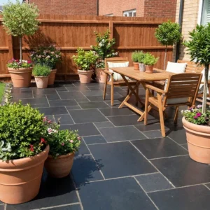 A patio paved with Black Limestone Sawn Edge - 22mm Calibrated Mixed Sizes, featuring a wooden table, chairs, large terracotta pots with lush plants and flowers, bordered by a wooden fence and a brick wall.