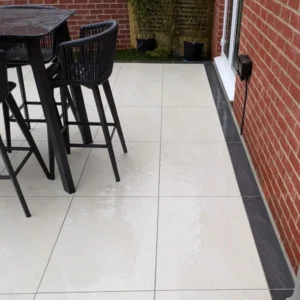 A modern patio with County Ivory Outdoor Porcelain Paving Tiles (600mm x 900mm), black border, tall black table and chairs, set by a red brick wall and glass door. Bamboo plants add greenery in the corner.