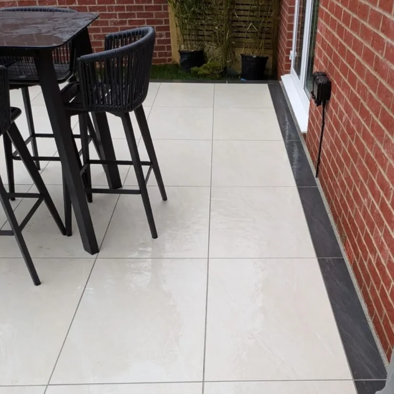A modern patio with County Ivory Outdoor Porcelain Paving Tiles (600mm x 900mm), black border, tall black table and chairs, set by a red brick wall and glass door. Bamboo plants add greenery in the corner.