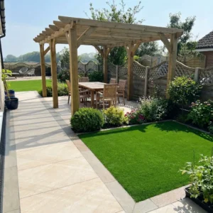 A backyard patio with a wooden pergola over a dining set, bordered by green grass, potted plants, flower beds, and wooden fencing features County Crema Outdoor Porcelain Paving Tiles (600mm x 900mm x 20mm) on the walkway.