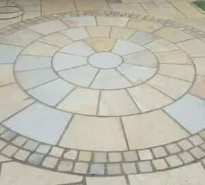 A circular patio design features the Yellow Indian Limestone 3 Meter Circle, showcasing concentric rings crafted from rectangular and square tiles and bordered by cobble setts. The pattern sits on a larger Indian sandstone paving surface, with grass visible in the corner.