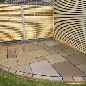 A patio featuring large rectangular and square Raj Green Sandstone tiles is bordered by a strip of smaller bricks. It is adjacent to a wooden slatted wall and fence, with a grassy area visible in the foreground. The sandstone tiles display varying shades of brown and beige.