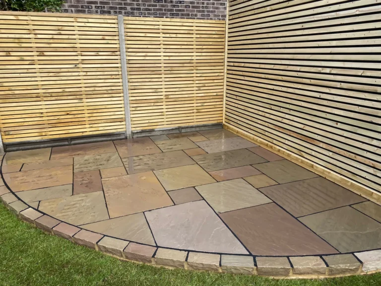 A patio featuring large rectangular and square Raj Green Sandstone tiles is bordered by a strip of smaller bricks. It is adjacent to a wooden slatted wall and fence, with a grassy area visible in the foreground. The sandstone tiles display varying shades of brown and beige.