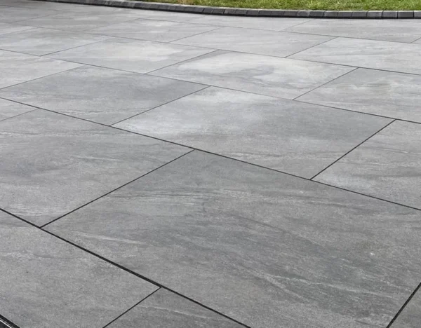 Close-up of a smooth, expansive surface featuring large rectangular tiles from the County Gris Outdoor Porcelain Paving sample collection. The natural-looking pavers are meticulously organized in a grid pattern and framed by a strip of lush green grass in the background.