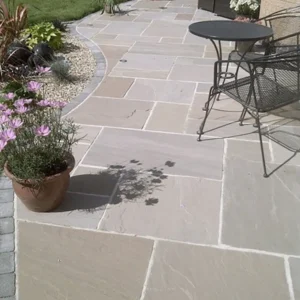 A stone patio with Raj Green - 22mm Calibrated Natural Mixed Size tiles showcases a black metal chair and round table. Nearby, a potted plant with purple flowers adds a splash of color, while the edge of a garden with grass and gravel is visible in the background.