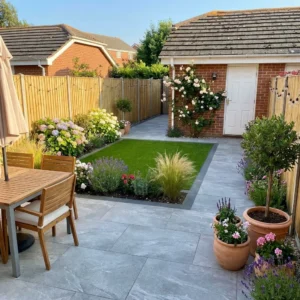 A neatly landscaped backyard features Earthcore Grey Outdoor Porcelain Paving Tiles (600x900mm), a small green lawn, flower beds, potted plants, a wooden patio table with chairs, a brick shed with a white door, and is enclosed by a wooden fence.