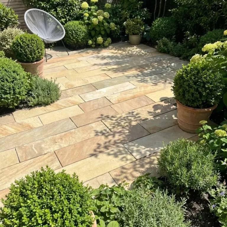 A sunlit garden patio features Fossil Mint Indian Sandstone Paving Planks (900x200x22mm), potted boxwood shrubs, lush plants, blooming hydrangeas, a modern white chair in the corner, and dappled shadows from nearby foliage.