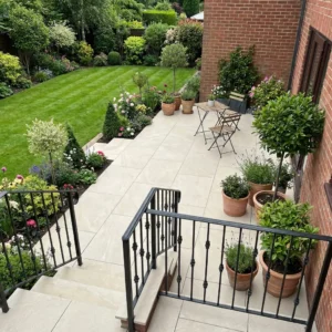 A view from above of a patio with potted plants, a small table with two chairs on Getafe Beige Porcelain paving, and stairs leading to a manicured garden with green grass, flowers, shrubs, and a brick house wall.