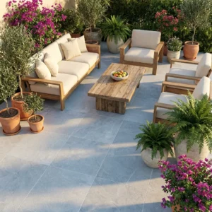 A cozy patio with wooden outdoor furniture, cream cushions, a wooden coffee table with a fruit bowl, and potted ferns, olive trees, and blooming flowers sits on Getafe Grey Outdoor Porcelain Paving Tiles (600×900×20mm).
