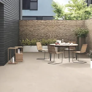 A contemporary patio showcases a dining set with four chairs and a table, placed on elegant Getafe Beige Outdoor Porcelain Paving Tiles. The space is enclosed by brick walls and decorated with potted plants, featuring sunny spots on the beige tiled floor. A glass door offers convenient access indoors.