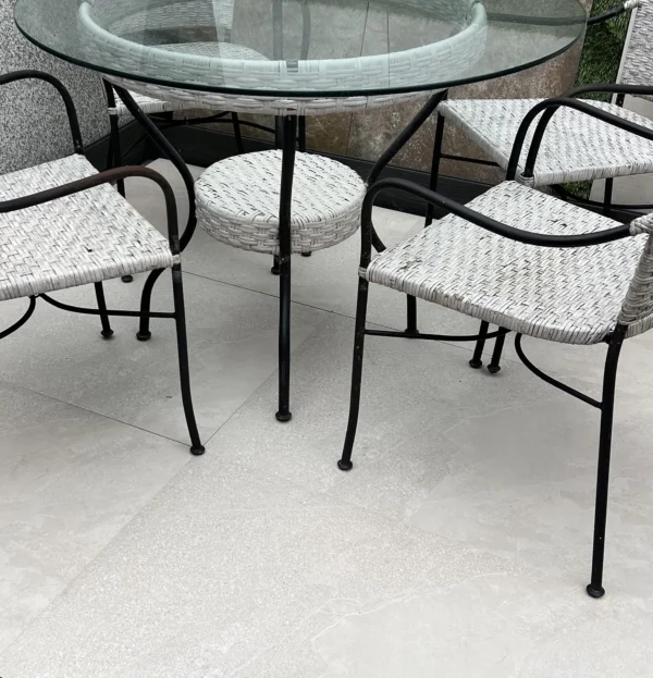 On a patio adorned with Getafe Beige Outdoor Porcelain Paving Tiles measuring 600mm by 900mm, an outdoor seating arrangement is featured with a round glass-top table surrounded by four woven chairs. These chairs have black metal frames and light-colored wicker seats, enhancing the rustic appeal of the porcelain paving underfoot.