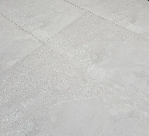 Close-up of the Getafe Beige Outdoor Porcelain Paving Tiles in light gray, featuring a textured surface with subtle patterns. The tiles have visible seams at their joints, forming a geometric design. The slightly glossy finish adds a gentle reflection to the surface.