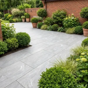 A modern garden patio features Grey Anthracite Outdoor Porcelain Paving Tiles (600mm x 900mm x 20mm), bordered by neatly trimmed shrubs, potted plants, and lush greenery set against a red brick wall.
