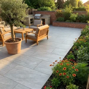 A modern patio features Grey Anthracite Outdoor Porcelain Paving Tiles (600mm x 600mm x 20mm), paired with wooden furniture, a potted tree, built-in barbecue grill, vibrant garden flowers, and lush greenery.