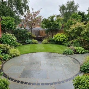 A lush, green garden features a circular patio of Grey Limestone Patio Paving - Sawn Edge - 22mm Calibrated - Mixed Size, bordered by black bricks and surrounded by trimmed grass, shrubs, flowers, trees, and wooden fences.