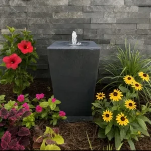 A small, modern black stone fountain stands amid red hibiscus, pink petunias, and yellow black-eyed Susans, set against a gray wall clad in Jak Black Splitface Stone Cladding.