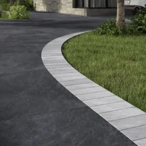 Curved driveway with large Kandla Grey Outdoor Porcelain Cobble Setts, bordered by light tiles, next to a trimmed green lawn and trees, in front of a modern stone building.