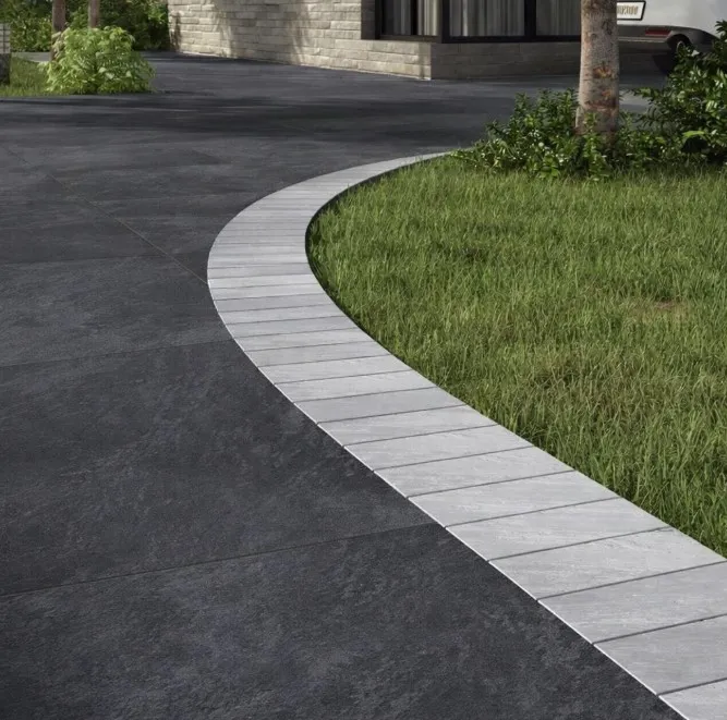 Curved driveway with large Kandla Grey Outdoor Porcelain Cobble Setts, bordered by light tiles, next to a trimmed green lawn and trees, in front of a modern stone building.