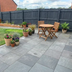 A patio paved with Kandla Grey Indian Sandstone Paving Slabs (22mm Calibrated, 600mm x 600mm) features a small round wooden table, two chairs, several potted plants along the edge, a dark wooden fence, and grass in the background.