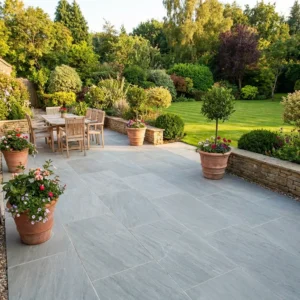 A spacious patio paved with Kandla Grey Sandstone Sawn Edge slabs features a wooden dining set, surrounded by potted plants and colorful flowers, overlooking a lush garden with manicured shrubs and trees.