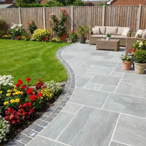 A patio paved with Kandla Grey Sandstone - 22mm Calibrated Mixed Size showcases potted plants and wicker furniture, curving into a green lawn bordered by colorful flowers, with a wooden fence and houses behind.