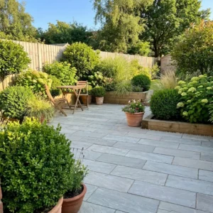 A sunny patio garden with Kandla Grey Indian Sandstone Paving Planks, a wooden table and two chairs, lush green plants, shrubs, potted flowers, all enclosed by a wooden fence.