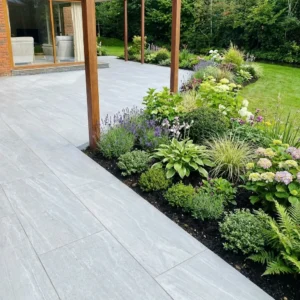 A modern patio features Kandla Grey Outdoor Porcelain Paving Tiles (600x900x20mm) beside a garden bed of green plants and flowers, bordered by wooden posts, with a lawn and trees in the background.