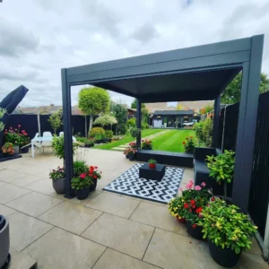 Modern garden patio with a pergola, potted plants, and a black-and-white rug on Mocha Outdoor Porcelain Paving Tiles (600mm x 900mm x 20mm); lush lawn and greenery visible in the background beneath a cloudy sky.