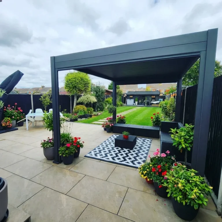 Modern garden patio with a pergola, potted plants, and a black-and-white rug on Mocha Outdoor Porcelain Paving Tiles (600mm x 900mm x 20mm); lush lawn and greenery visible in the background beneath a cloudy sky.