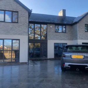 Modern two-story house with large windows and an exterior finished in Oriental White Cladding. A gray Range Rover is parked on the paved driveway, a trailer is visible to the right, and trees are reflected in the windows.
