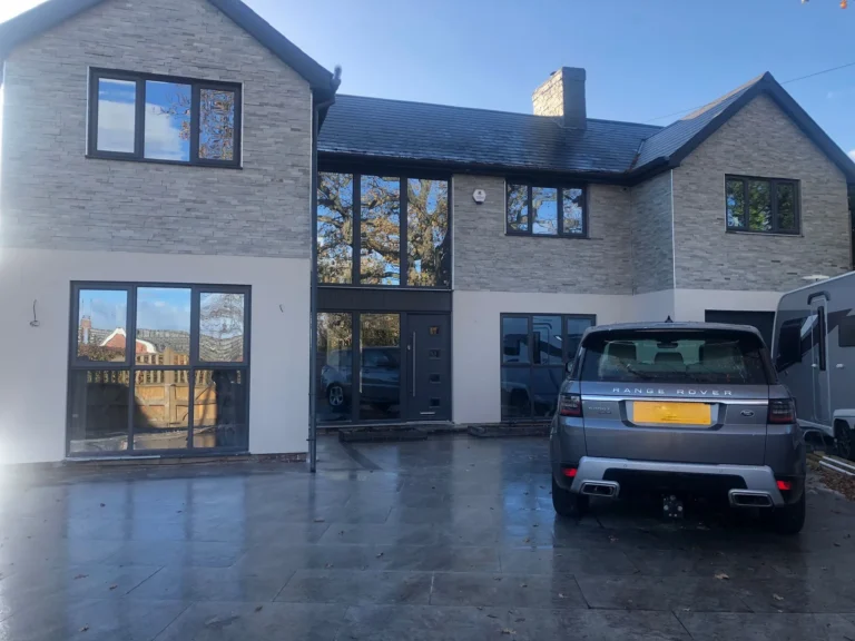 Modern two-story house with large windows and an exterior finished in Oriental White Cladding. A gray Range Rover is parked on the paved driveway, a trailer is visible to the right, and trees are reflected in the windows.