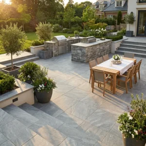 Spacious backyard patio with stone tiles, an outdoor dining table set with a white cloth, two built-in grills with stone surrounds, potted plants, and lush green landscaping in the background.